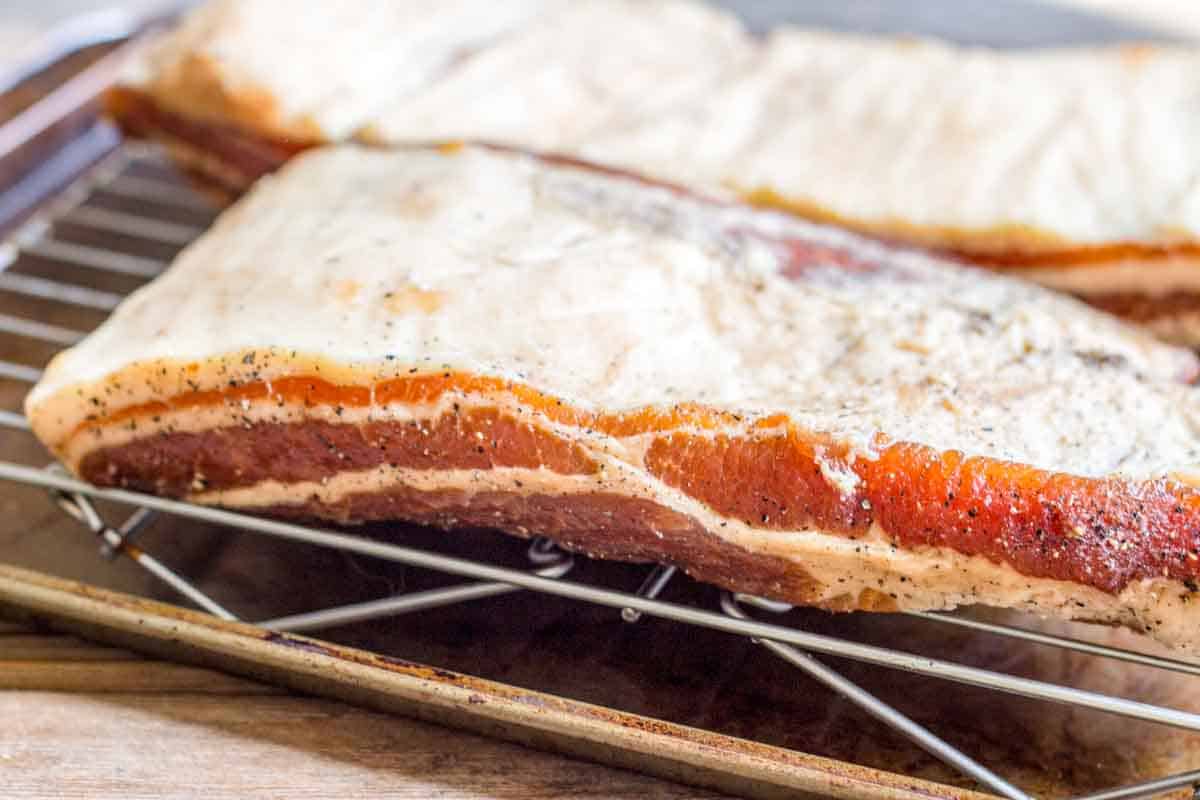 Close-up of raw bacon slabs on a wire rack showing the cross-section thickness. The bacon displays a generous fat cap with pinkish-red meat and black pepper seasoning, ready for smoking or further preparation.
