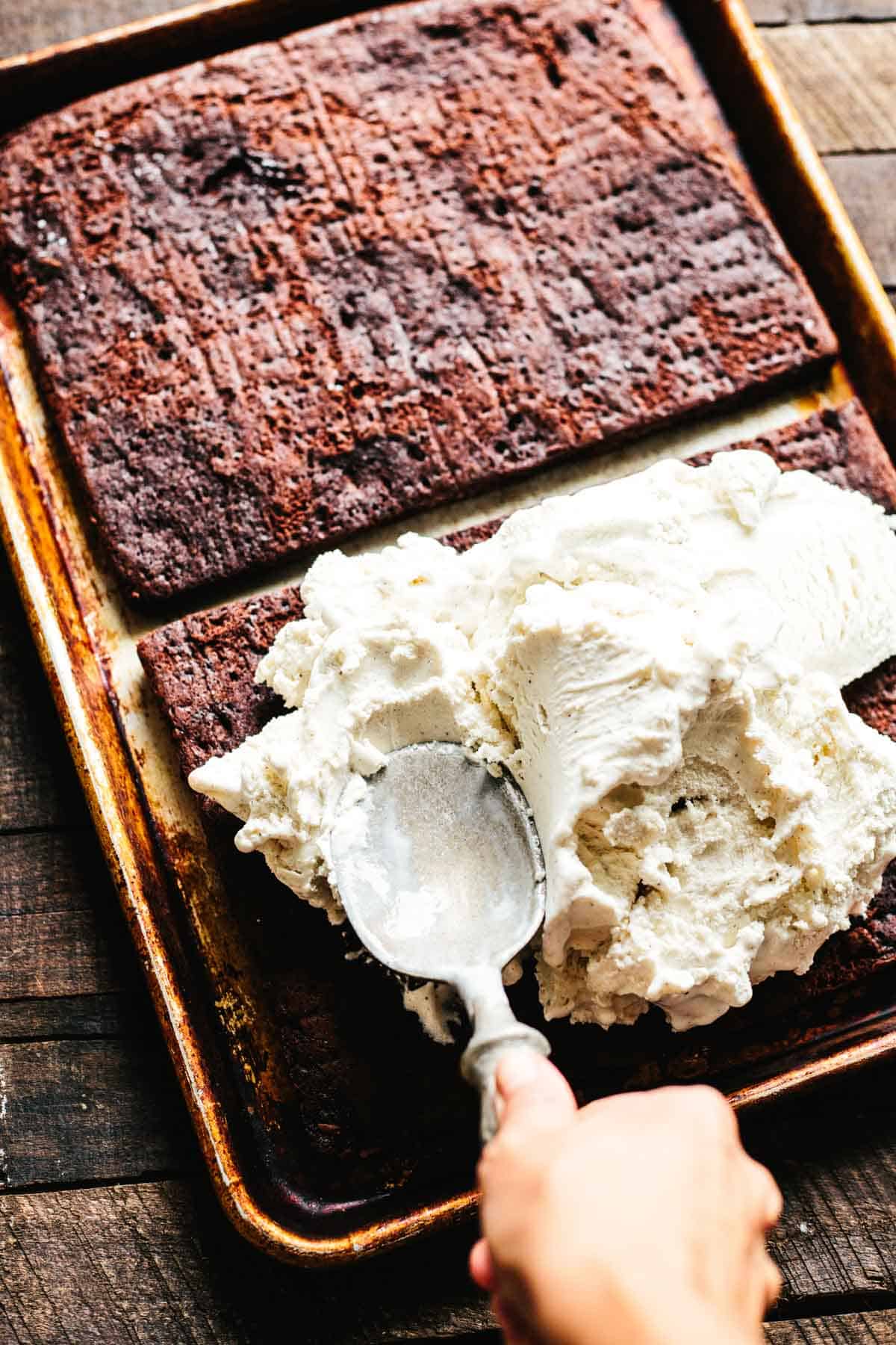 A hand uses an old-fashioned metal ice cream scoop to spread a thick layer of vanilla ice cream onto a large, rectangular sheet of soft chocolate cookie, set inside a rimmed baking sheet.