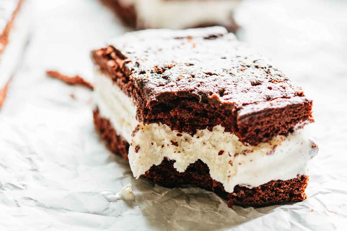 A close-up of a partially eaten ice cream sandwich resting on parchment paper, with a visible bite revealing creamy vanilla ice cream inside thick soft chocolate cookie layers.