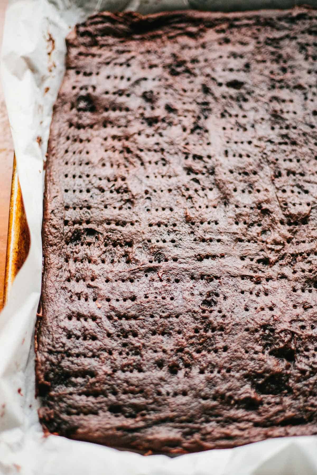 A single large sheet of baked soft chocolate cookie pierced with fork marks, still in a parchment-lined baking pan, ready to be layered or sliced.