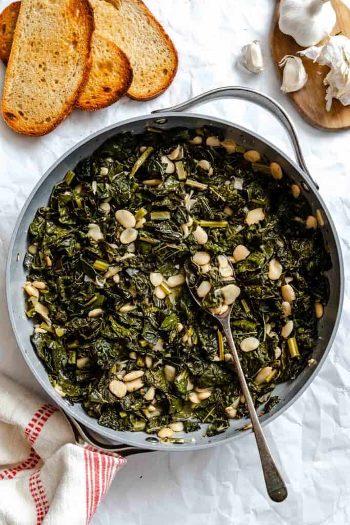 Finished Tuscan kale and beans dish served in a large skillet, with toasted bread slices and garlic cloves on a wooden board alongside. A metal spoon rests in the skillet, showing the moist, saucy consistency of the greens and beans.