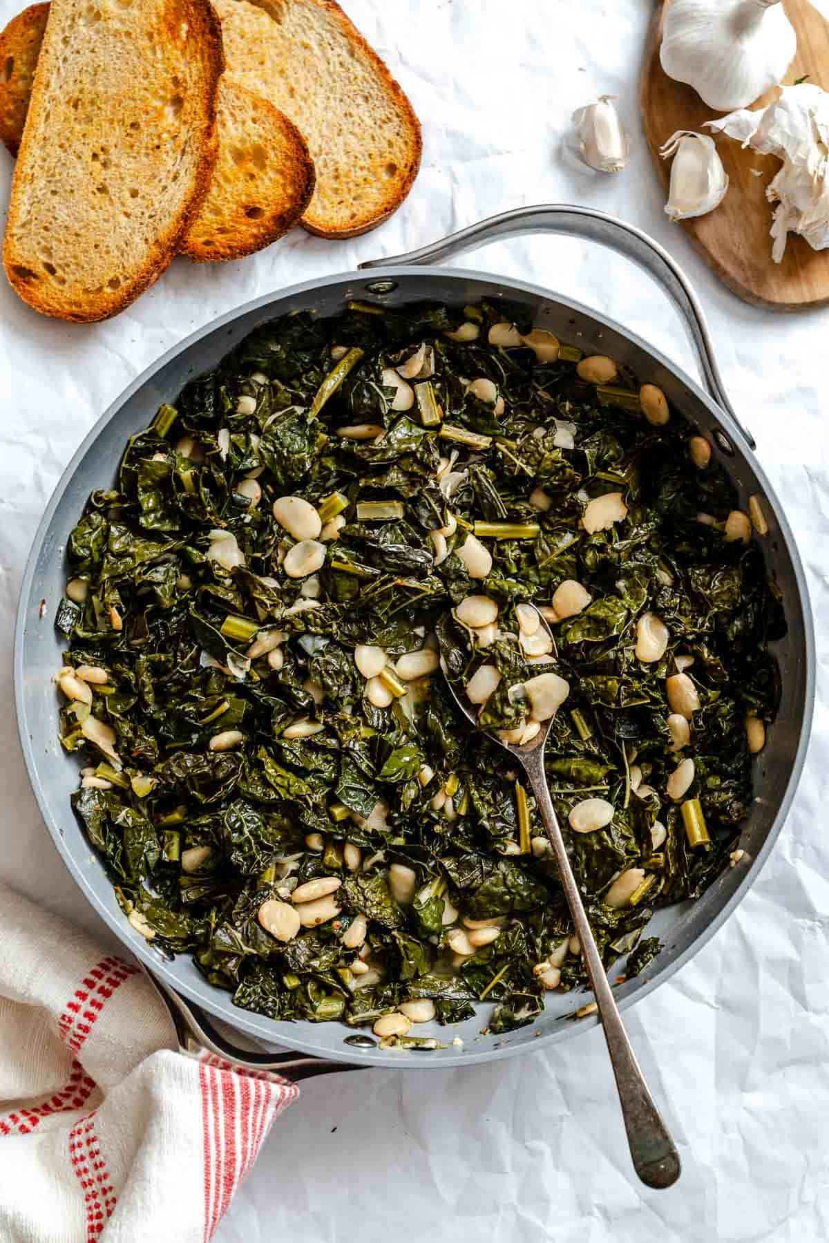 Finished Tuscan kale and beans dish served in a large skillet, with toasted bread slices and garlic cloves on a wooden board alongside. A metal spoon rests in the skillet, showing the moist, saucy consistency of the greens and beans.