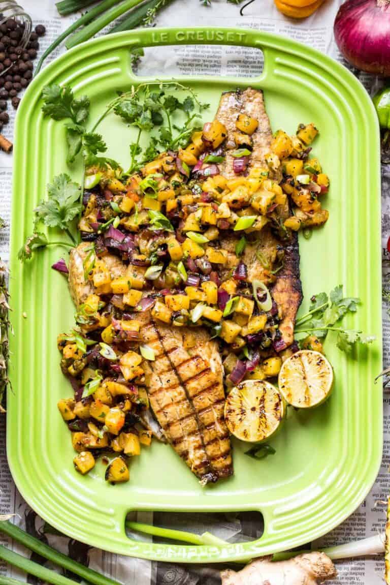 Grilled fish fillet on a bright lime-green Le Creuset platter, topped with vibrant yellow pineapple salsa featuring red onions, green scallions, and fresh cilantro. Charred lime halves accompany the dish, which is presented on newspaper.