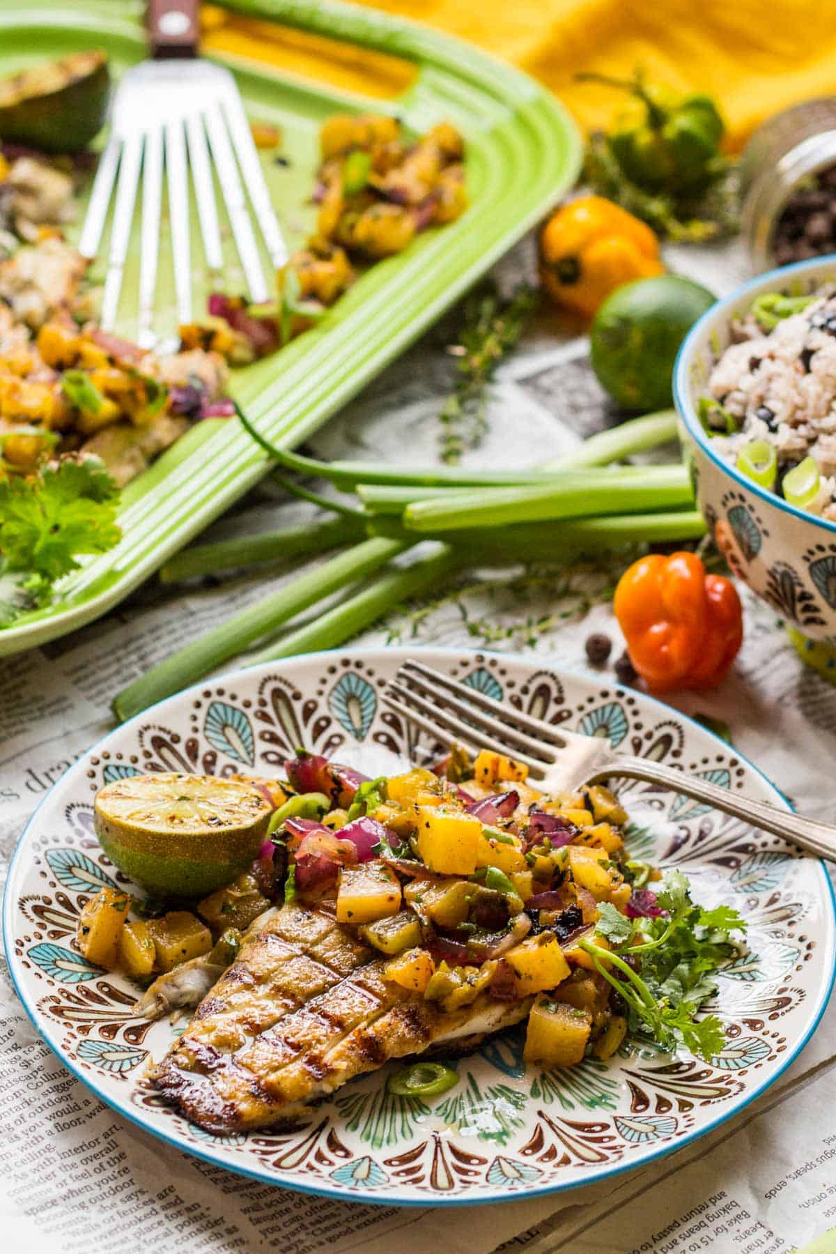 Individual serving of jerk-seasoned fish on a decorative plate with blue rim and intricate pattern, generously covered with colorful mango-pineapple salsa. Rice in a small bowl and fresh ingredients are scattered artfully around the rustic newspaper setting.