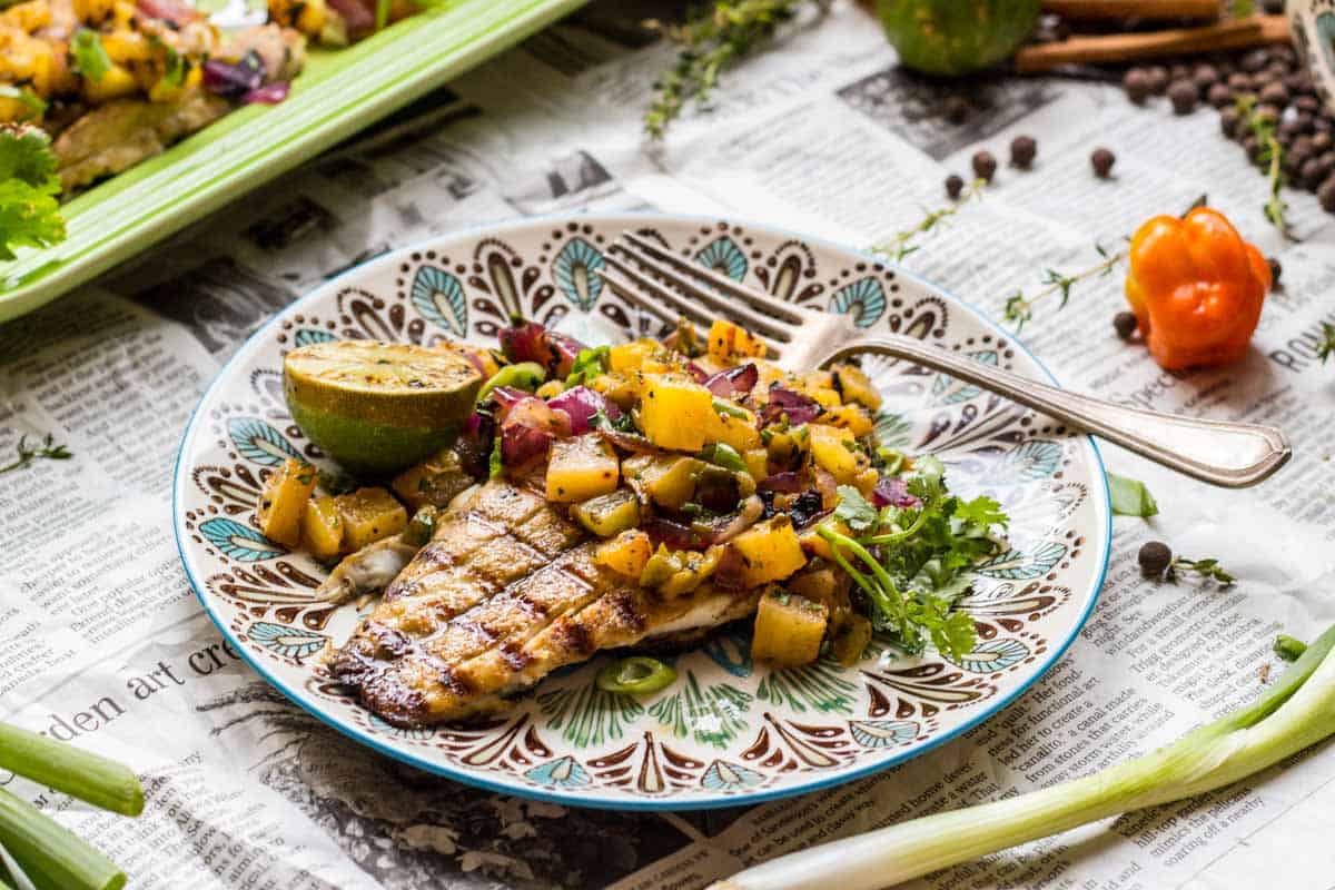 Close-up of jerk fish with tropical salsa on an ornate plate, showcasing the perfectly grilled fish beneath chunks of golden pineapple, purple onion, and bright herbs. Habanero pepper and spices visible in the rustic backdrop.