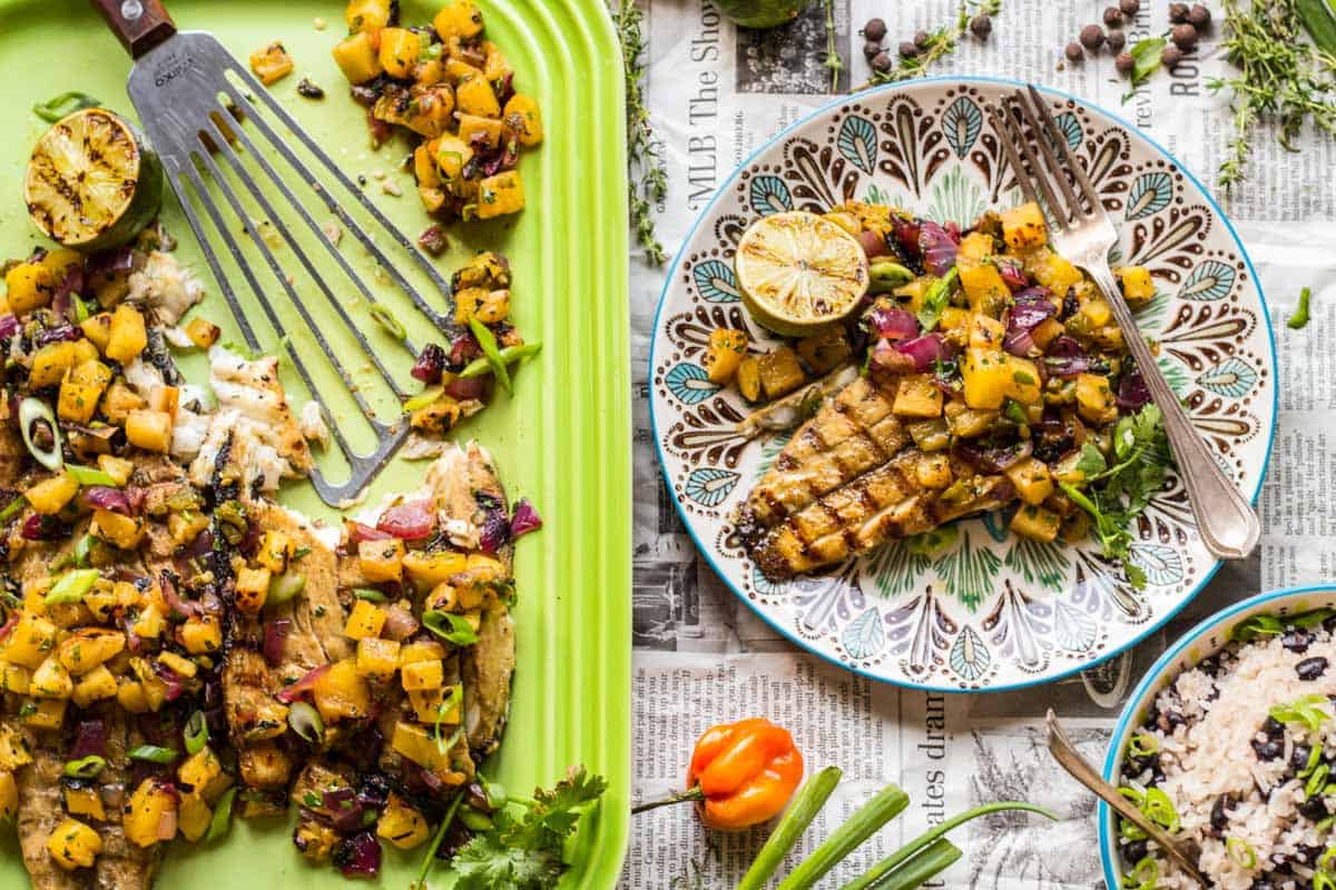 Side-by-side presentation showing fish on both the green Le Creuset platter with visible fork marks and an individual decorative plate, emphasizing the contrast between family-style and single serving presentations of the same Caribbean-inspired dish.