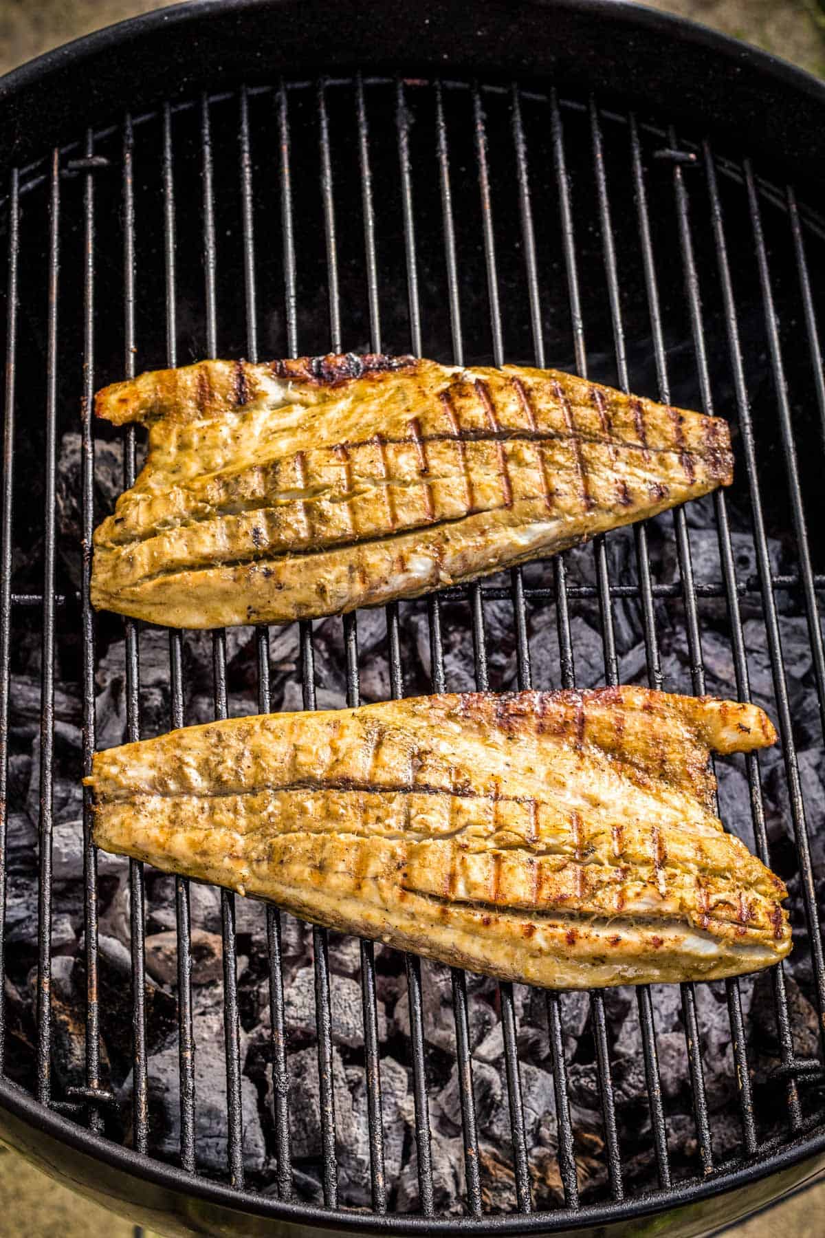 Two fish fillets cooking directly on a round grill, displaying distinctive cross-hatch grill marks and golden-brown jerk seasoning. The fillets are shown mid-cooking over hot coals, capturing the authentic grilling technique.