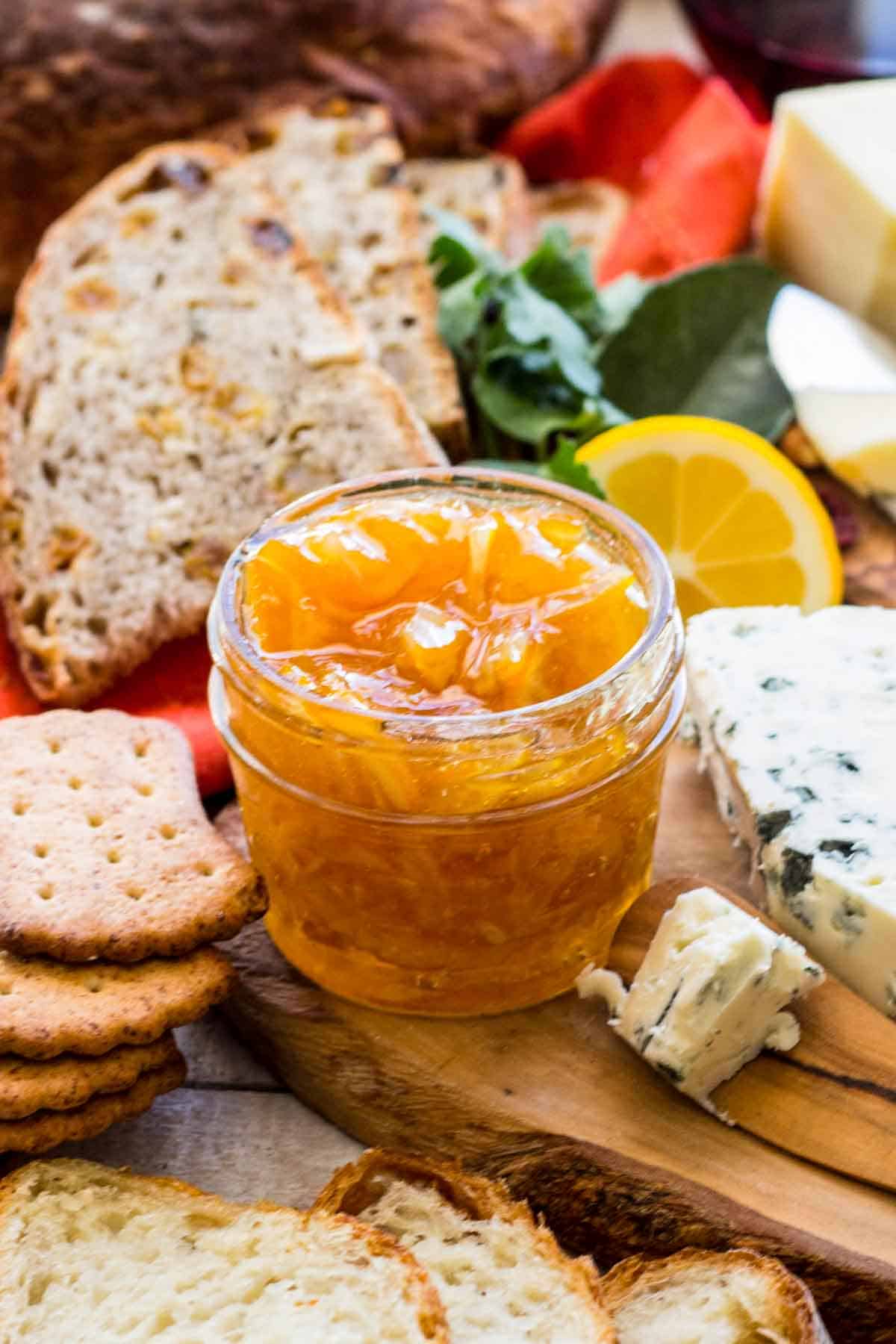 Meyer lemon marmalade in a glass container surrounded by crackers, brown bread, and different kinds of cheeses.