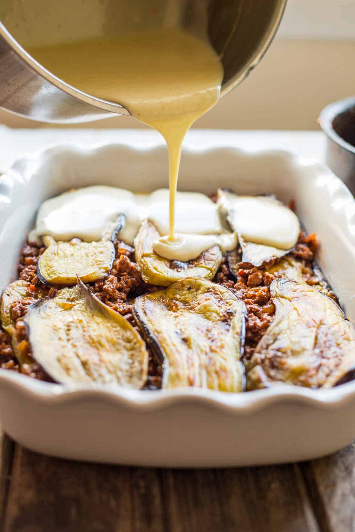 Golden béchamel sauce being poured from a metal pot onto a baking dish containing layers of roasted eggplant slices and seasoned ground meat mixture. The sauce creates an appetizing stream as it's added to complete the moussaka assembly.