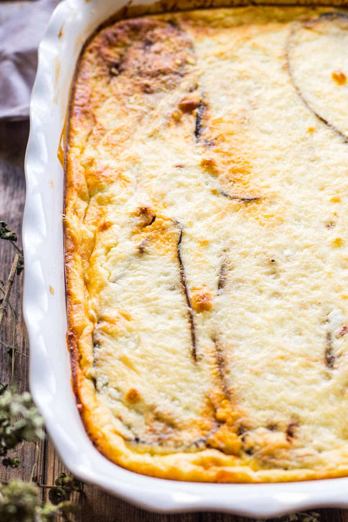 A freshly baked moussaka in a white ceramic baking dish with scalloped edges, showing the golden brown, slightly bubbling cheese topping. The creamy béchamel surface has browned perfectly with visible outlines of the eggplant layers beneath.