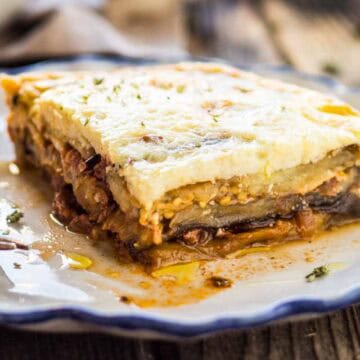 A single portion of moussaka on a decorative white and blue-rimmed plate. The slice reveals distinct layers of eggplant, meat sauce, and creamy béchamel topping, with juices from the dish spreading on the plate. A fork rests alongside, ready for tasting.