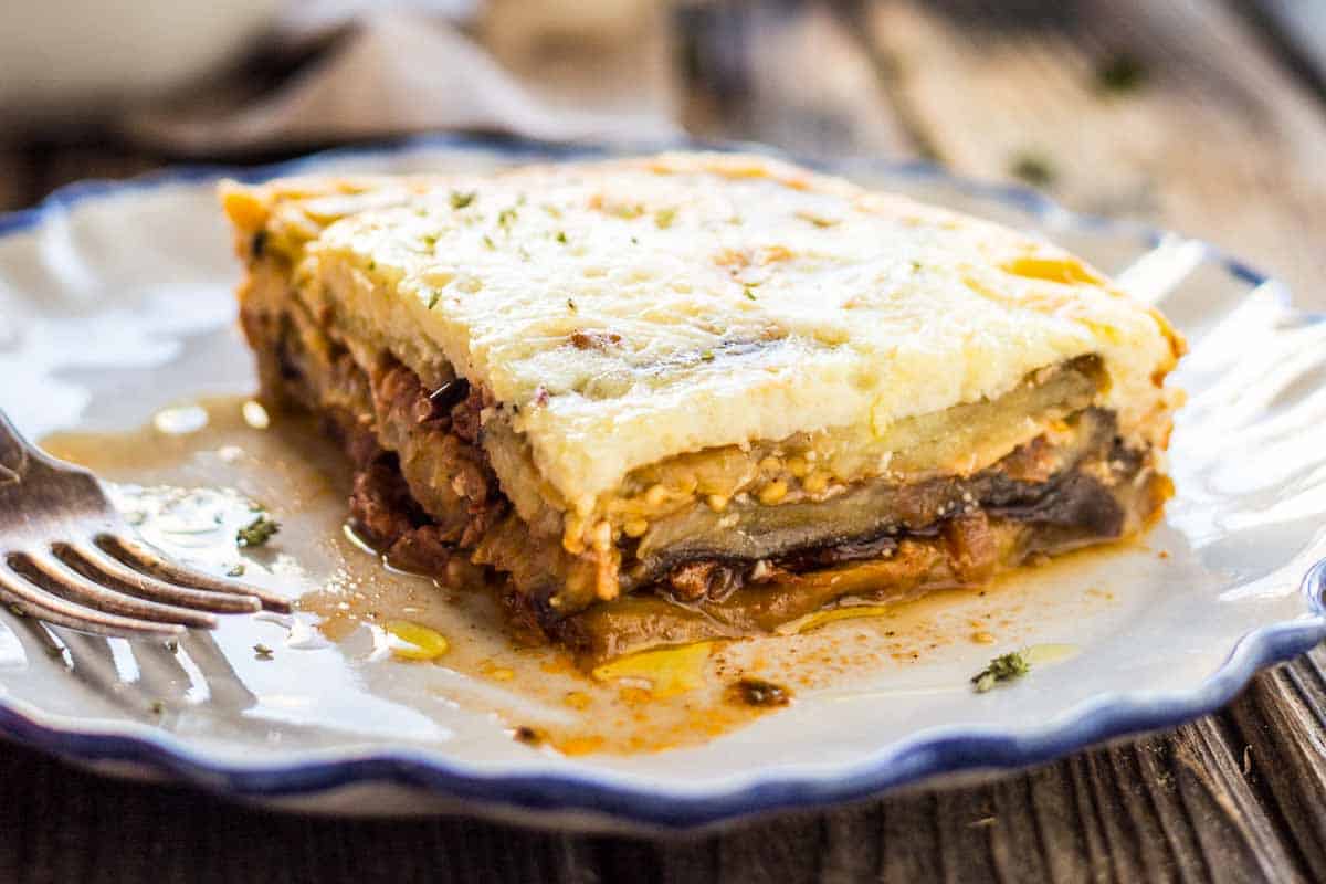 A single portion of moussaka on a decorative white and blue-rimmed plate. The slice reveals distinct layers of eggplant, meat sauce, and creamy béchamel topping, with juices from the dish spreading on the plate. A fork rests alongside, ready for tasting.