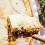 A serving of moussaka being lifted from the baking dish with a spatula. The image shows the perfect layering of the dish with its golden top, meaty middle layer, and eggplant base. The dish's sauces are visible bubbling around the edges of the baking dish.