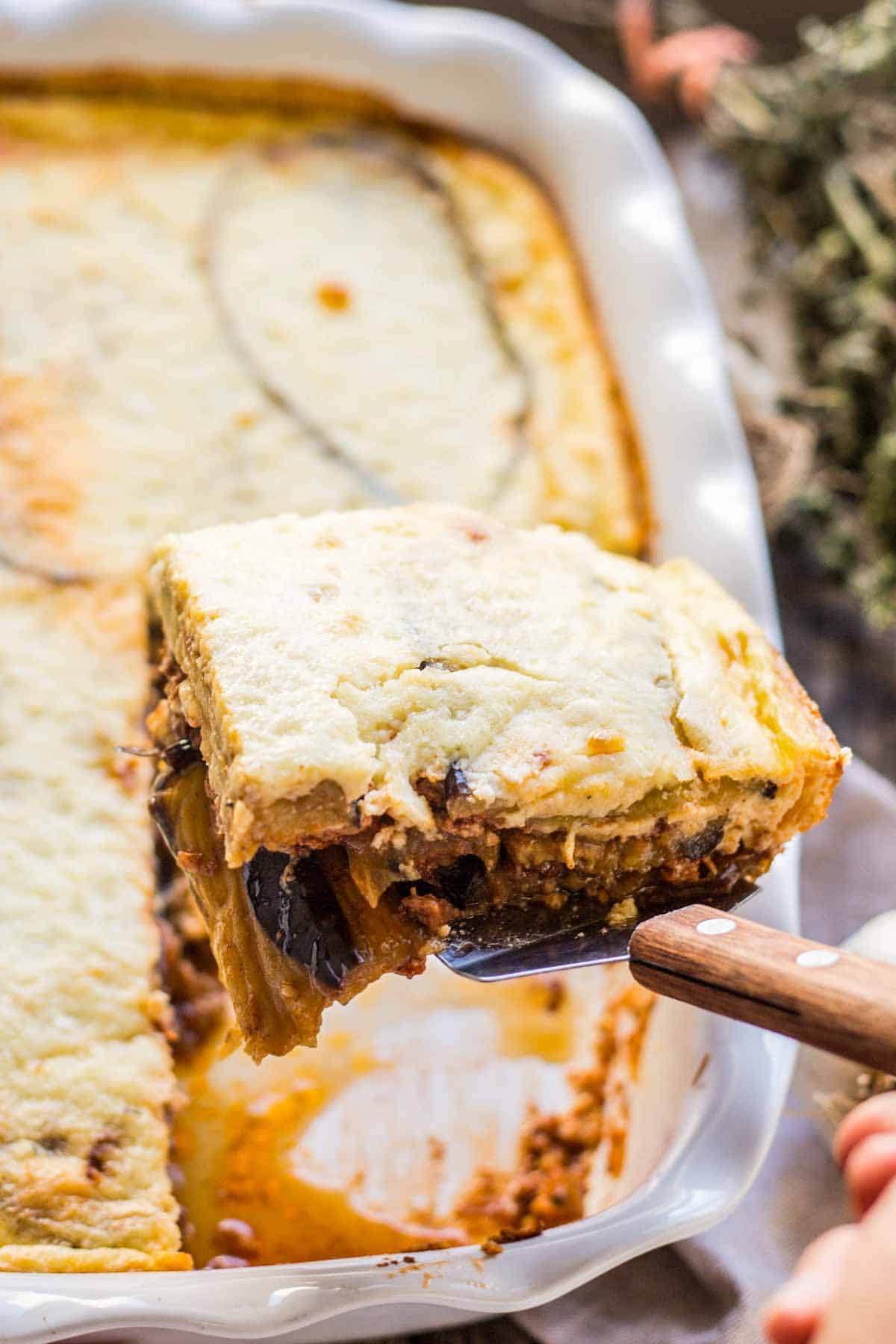 A serving of moussaka being lifted from the baking dish with a spatula. The image shows the perfect layering of the dish with its golden top, meaty middle layer, and eggplant base. The dish's sauces are visible bubbling around the edges of the baking dish.