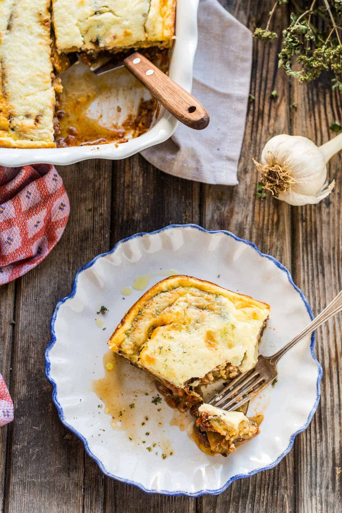 Overhead view of a complete moussaka serving setup, featuring the white ceramic baking dish with a section removed, a plated portion on a blue-rimmed plate with a bite taken, a kitchen towel, and fresh garlic bulb visible. The rustic wooden table adds warmth to the presentation.