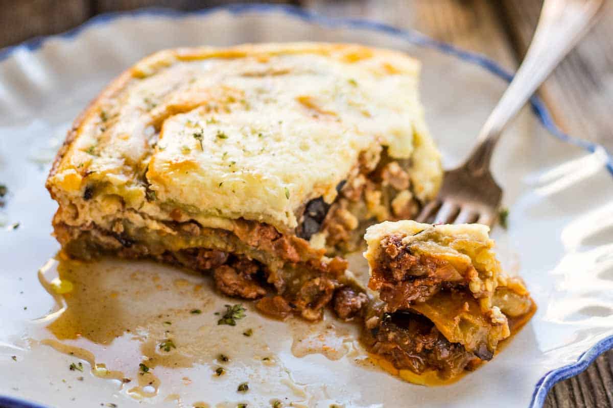 Close-up of a moussaka portion on a blue-rimmed plate. A forkful has been taken, revealing the distinct layers of creamy béchamel, seasoned ground meat, and tender eggplant. The plate has sauce pooling around the slice, with herbs sprinkled on top.