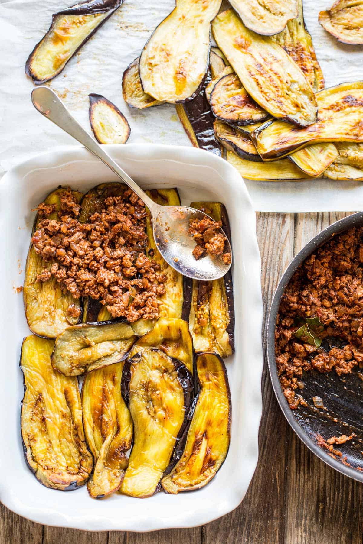 Preparation stage of moussaka showing two dishes - one white ceramic baking dish lined with roasted eggplant slices being topped with seasoned ground meat mixture, alongside another roasting tray of golden-brown roasted eggplant slices. A skillet of remaining meat sauce is visible at the bottom right.