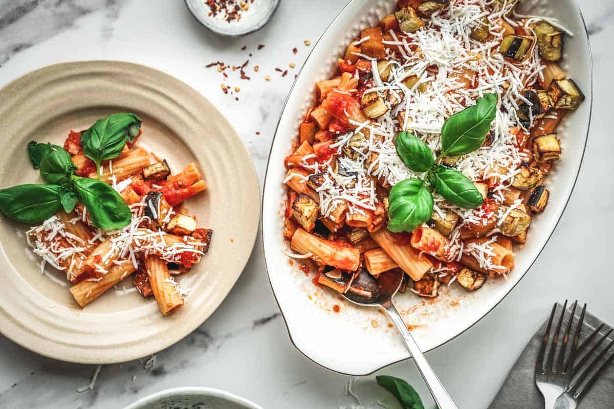A table setting with two portions of pasta alla norma - one in a small beige plate and another in the white oval serving dish. Both are garnished with basil and cheese, creating an inviting presentation for sharing.