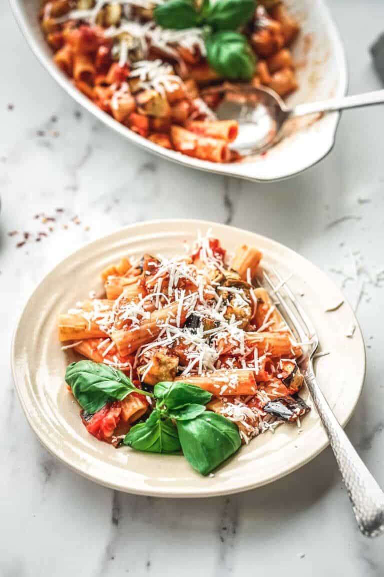 Close-up of an individual portion of pasta alla norma on a beige plate. The rigatoni is well-coated with sauce, dotted with eggplant pieces, sprinkled with cheese, and adorned with fresh basil. A silver fork rests on the plate, ready for dining.