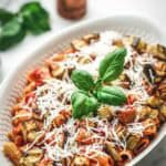 A closer view of the pasta alla norma in the serving dish, showing more detail of the perfectly coated rigatoni, roasted eggplant, and creamy ricotta salata topping. Fresh basil sprigs add color and a finishing touch.