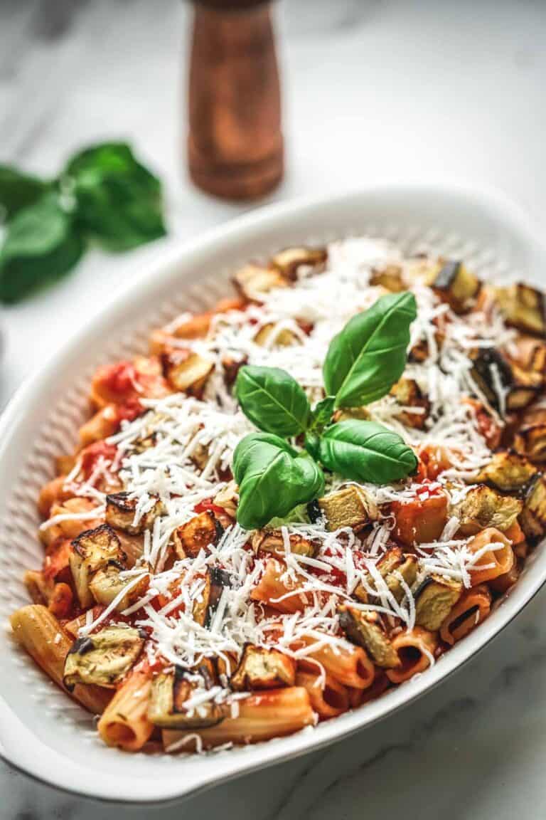 A closer view of the pasta alla norma in the serving dish, showing more detail of the perfectly coated rigatoni, roasted eggplant, and creamy ricotta salata topping. Fresh basil sprigs add color and a finishing touch.