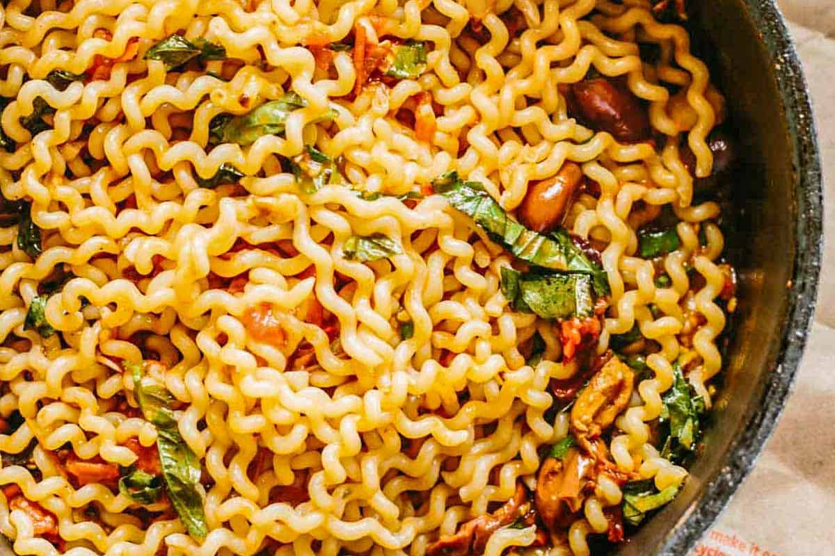 Close-up of a pan filled with wavy fusilli pasta tossed with a tomato-based sauce. The golden pasta is interspersed with bits of green basil, red tomatoes, and darker olives, creating a colorful and appetizing presentation.