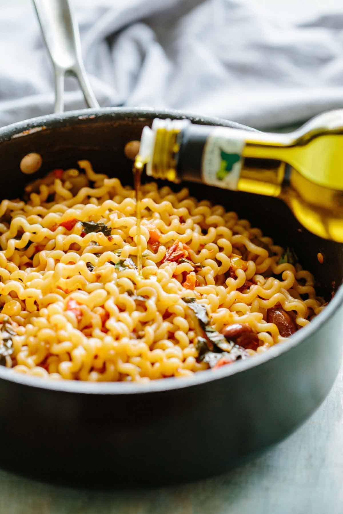 Olive oil being drizzled from a bottle onto fusilli pasta in a black skillet. The golden pasta is mixed with tomatoes, herbs, and olives, with the olive oil adding a finishing touch of richness and shine to the completed dish.