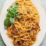 An elegant white oval serving platter featuring fusilli pasta topped with golden breadcrumbs and garnished with fresh basil leaves. The pasta is coated in a tomato-based sauce and has a rustic, homemade appearance against the teal-colored background.