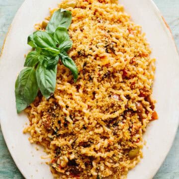 An elegant white oval serving platter featuring fusilli pasta topped with golden breadcrumbs and garnished with fresh basil leaves. The pasta is coated in a tomato-based sauce and has a rustic, homemade appearance against the teal-colored background.