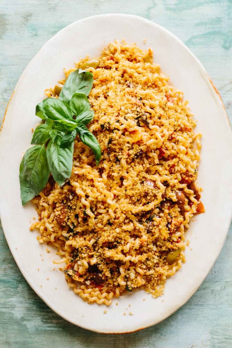 An elegant white oval serving platter featuring fusilli pasta topped with golden breadcrumbs and garnished with fresh basil leaves. The pasta is coated in a tomato-based sauce and has a rustic, homemade appearance against the teal-colored background.