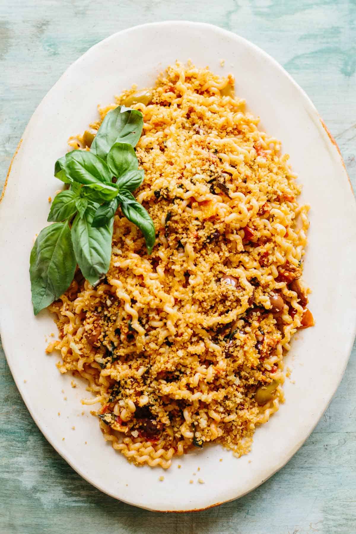 An elegant white oval serving platter featuring fusilli pasta topped with golden breadcrumbs and garnished with fresh basil leaves. The pasta is coated in a tomato-based sauce and has a rustic, homemade appearance against the teal-colored background.
