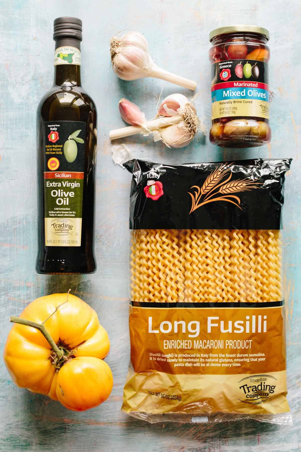 Ingredients for the pasta dish arranged on a teal surface: a bottle of Sicilian extra virgin olive oil, fresh garlic bulbs and cloves, a jar of mixed marinated olives, a package of long fusilli pasta, and a bright yellow tomato. These components form the base for the Mediterranean-inspired dish.