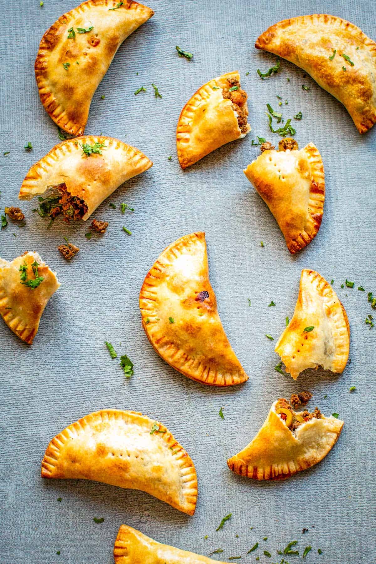 Golden-brown baked empanadas arranged on a light blue textured surface. The half-moon pastries have crimped edges and are garnished with chopped fresh herbs. Some are broken open, revealing the savory picadillo filling of seasoned ground beef with visible pieces of olives.