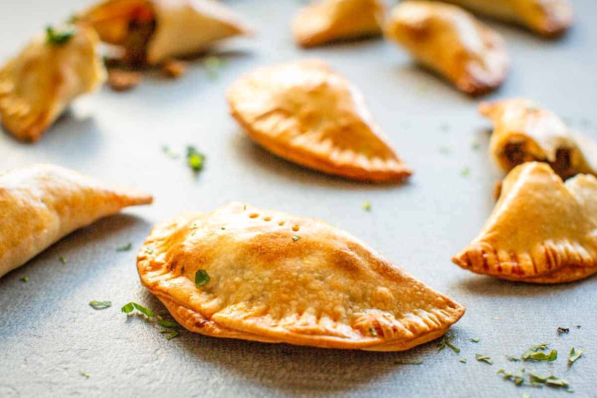 Close-up of freshly baked empanadas on a baking sheet. The empanadas have a perfect golden color with distinctive crimped edges and small steam vents. Scattered herbs add a touch of green against the flaky pastry.