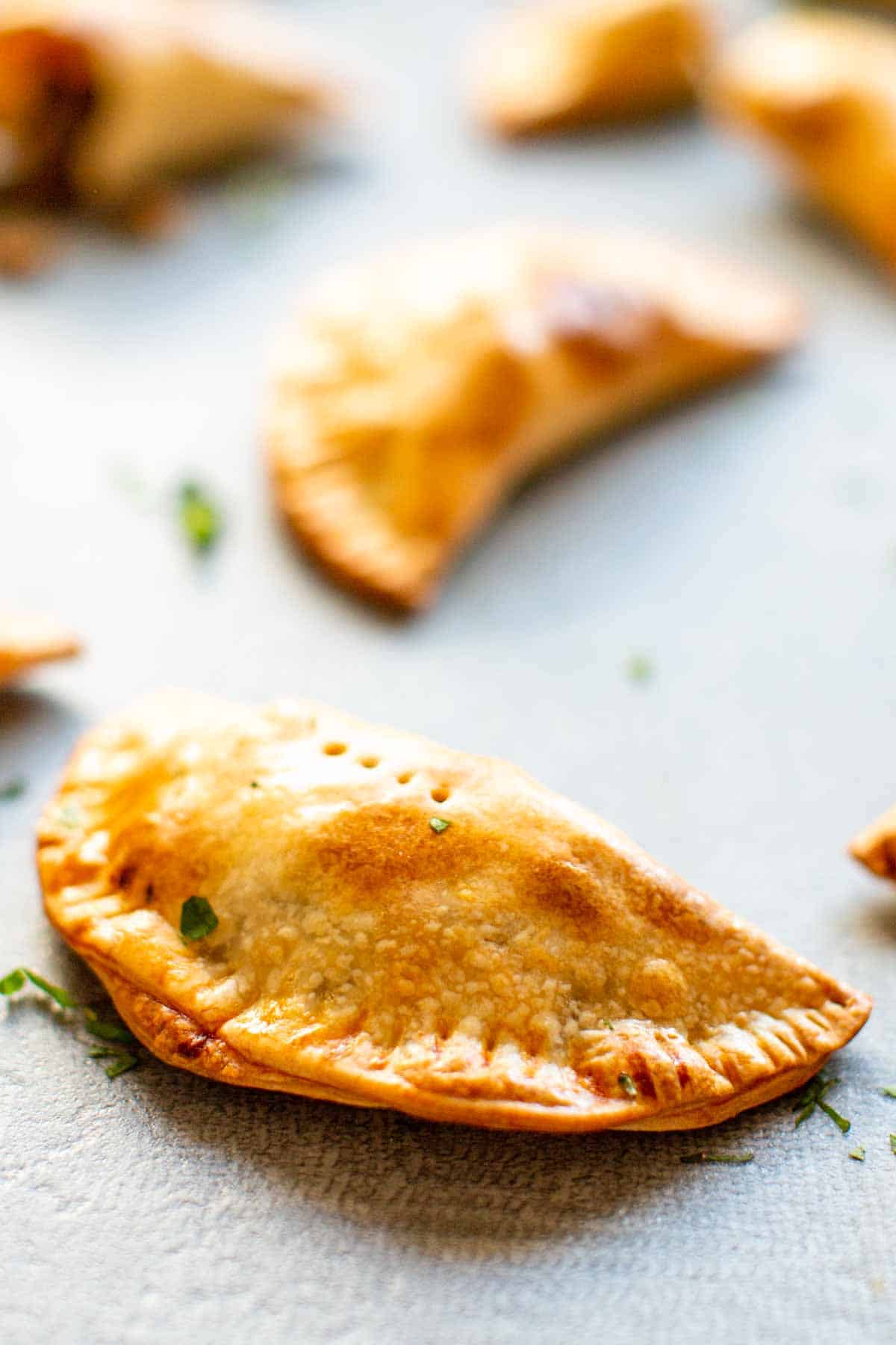 Extreme close-up of a single empanada showing the beautifully baked, flaky crust with small fork-made perforations. The pastry has an appetizing golden-brown color with tiny specks of herbs visible against the light-colored baking surface.