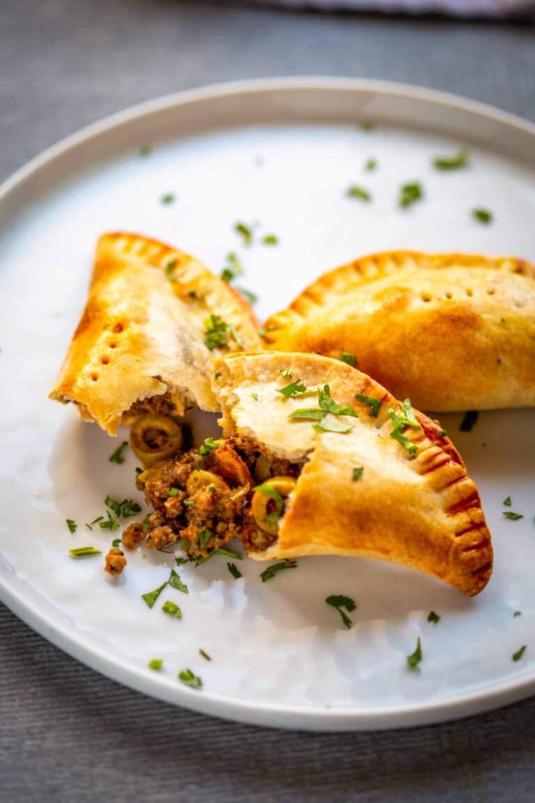 Three empanadas arranged on a white plate with one broken open to showcase the picadillo filling. The ground beef mixture contains visible pieces of olives and is garnished with fresh herbs. The empanada's flaky crust provides a perfect contrast to the savory filling.