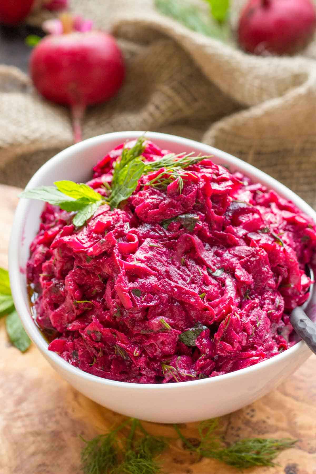 Close-up of roasted beet salad with visible herb flecks throughout the bright fuchsia mixture. Whole fresh beets appear in the blurred background along with burlap fabric, emphasizing the rustic, farm-to-table nature of the dish. Fresh mint and dill garnish the top, with the creamy dressing giving the salad a glossy appearance.