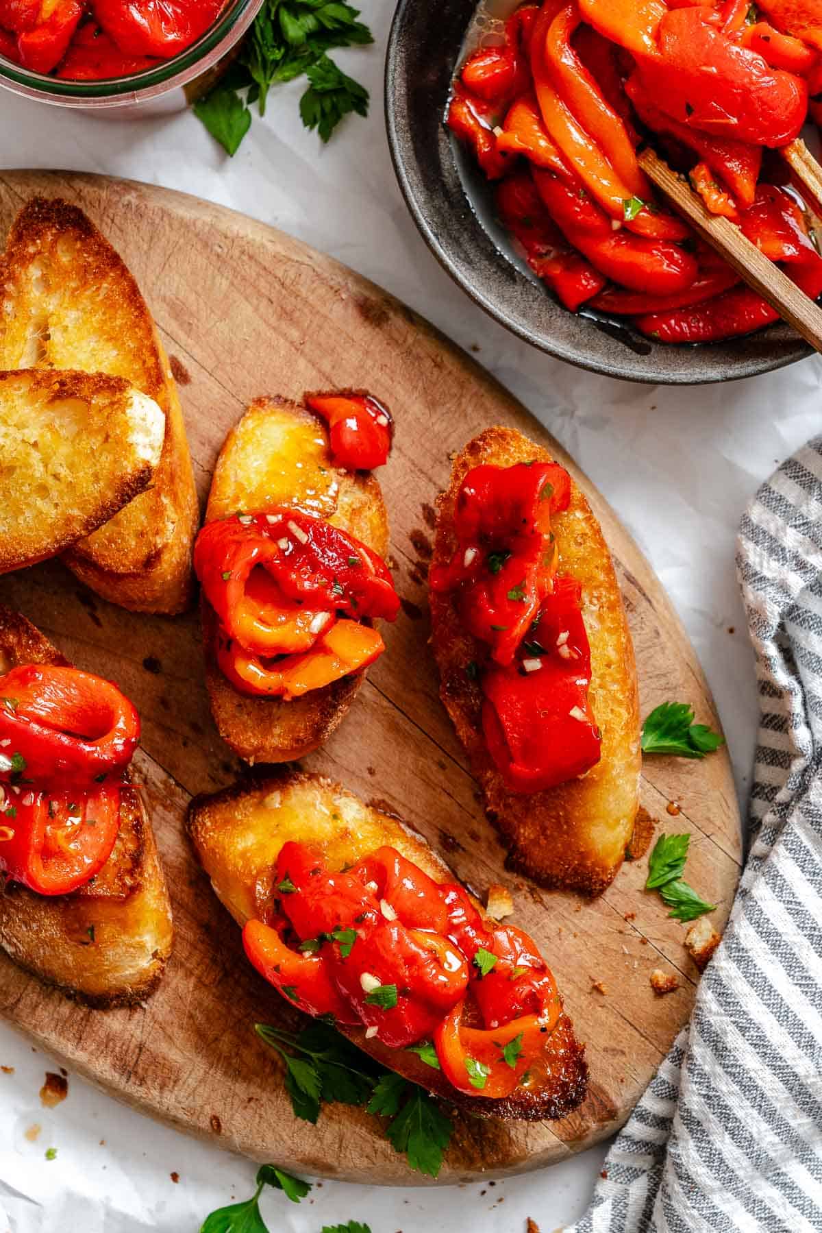 Toasted slices of crusty bread topped with marinated roasted red peppers, garnished with chopped herbs, arranged on a wooden board next to a bowl of extra peppers.