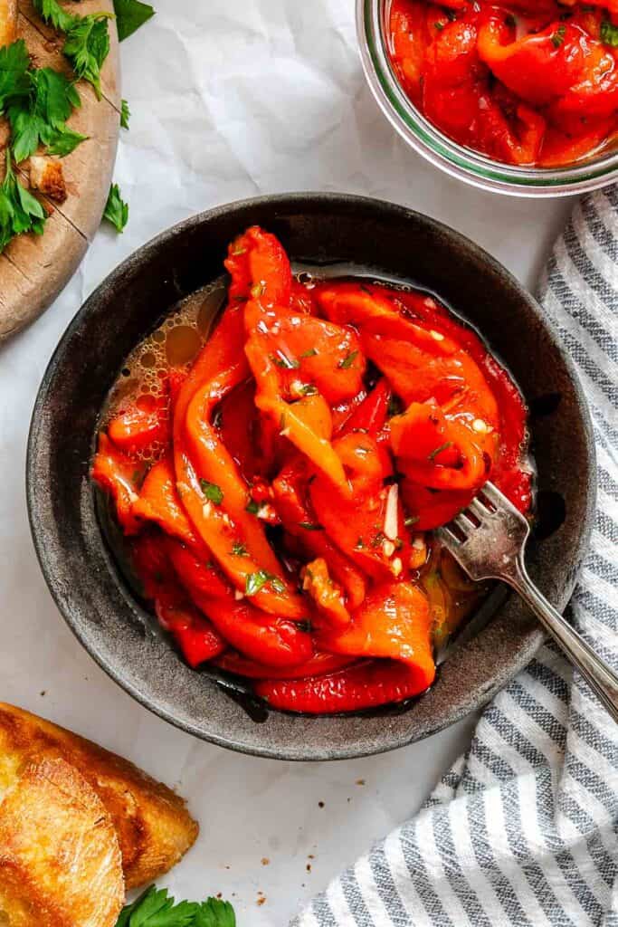 Close-up shot of a dark ceramic bowl filled with roasted red pepper strips marinated with olive oil, garlic, and herbs, with a fork resting inside the bowl.