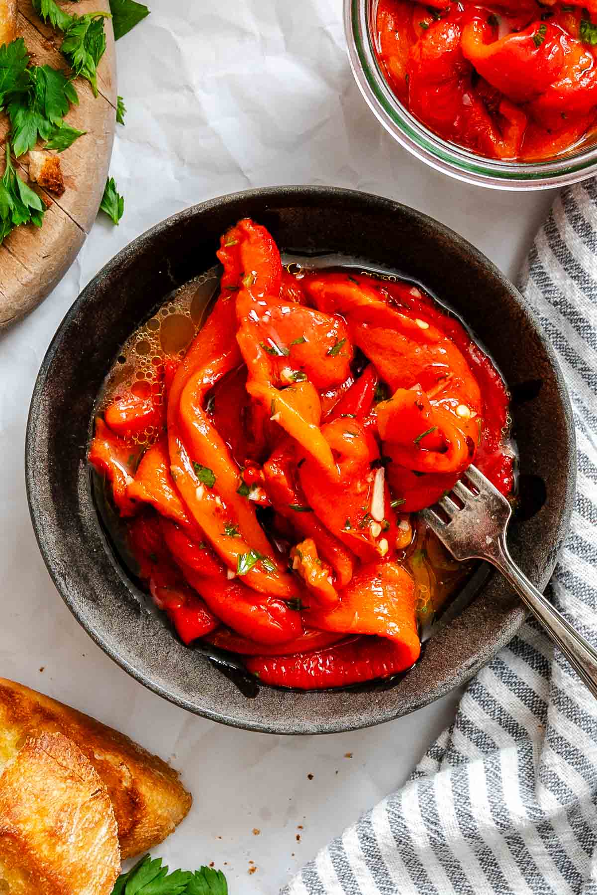 Close-up shot of a dark ceramic bowl filled with roasted red pepper strips marinated with olive oil, garlic, and herbs, with a fork resting inside the bowl.