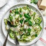 Finished salad in white speckled bowl garnished with shaved parmesan, chopped hazelnuts, and fresh basil. Surrounding ingredients include a block of parmesan, small bowl of hazelnuts, and fresh basil sprigs on white parchment background.