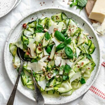 Finished salad in white speckled bowl garnished with shaved parmesan, chopped hazelnuts, and fresh basil. Surrounding ingredients include a block of parmesan, small bowl of hazelnuts, and fresh basil sprigs on white parchment background.