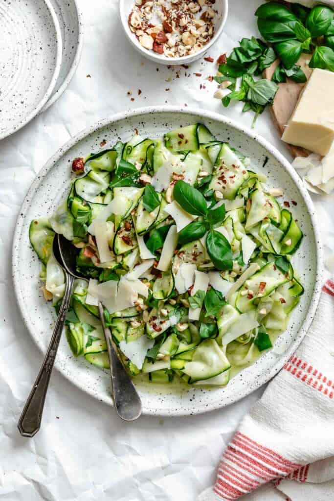 Finished salad in white speckled bowl garnished with shaved parmesan, chopped hazelnuts, and fresh basil. Surrounding ingredients include a block of parmesan, small bowl of hazelnuts, and fresh basil sprigs on white parchment background.