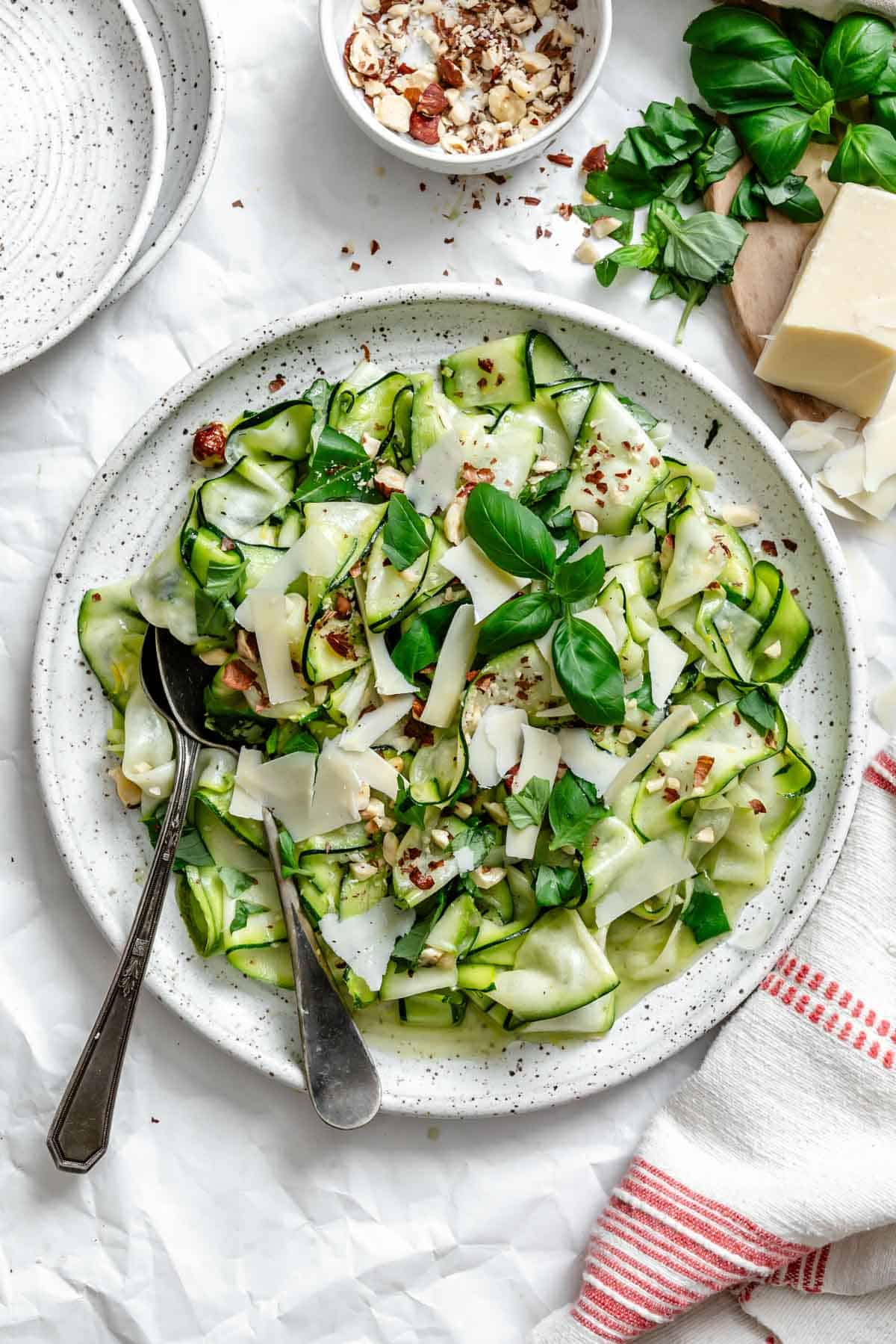 Finished salad in white speckled bowl garnished with shaved parmesan, chopped hazelnuts, and fresh basil. Surrounding ingredients include a block of parmesan, small bowl of hazelnuts, and fresh basil sprigs on white parchment background.