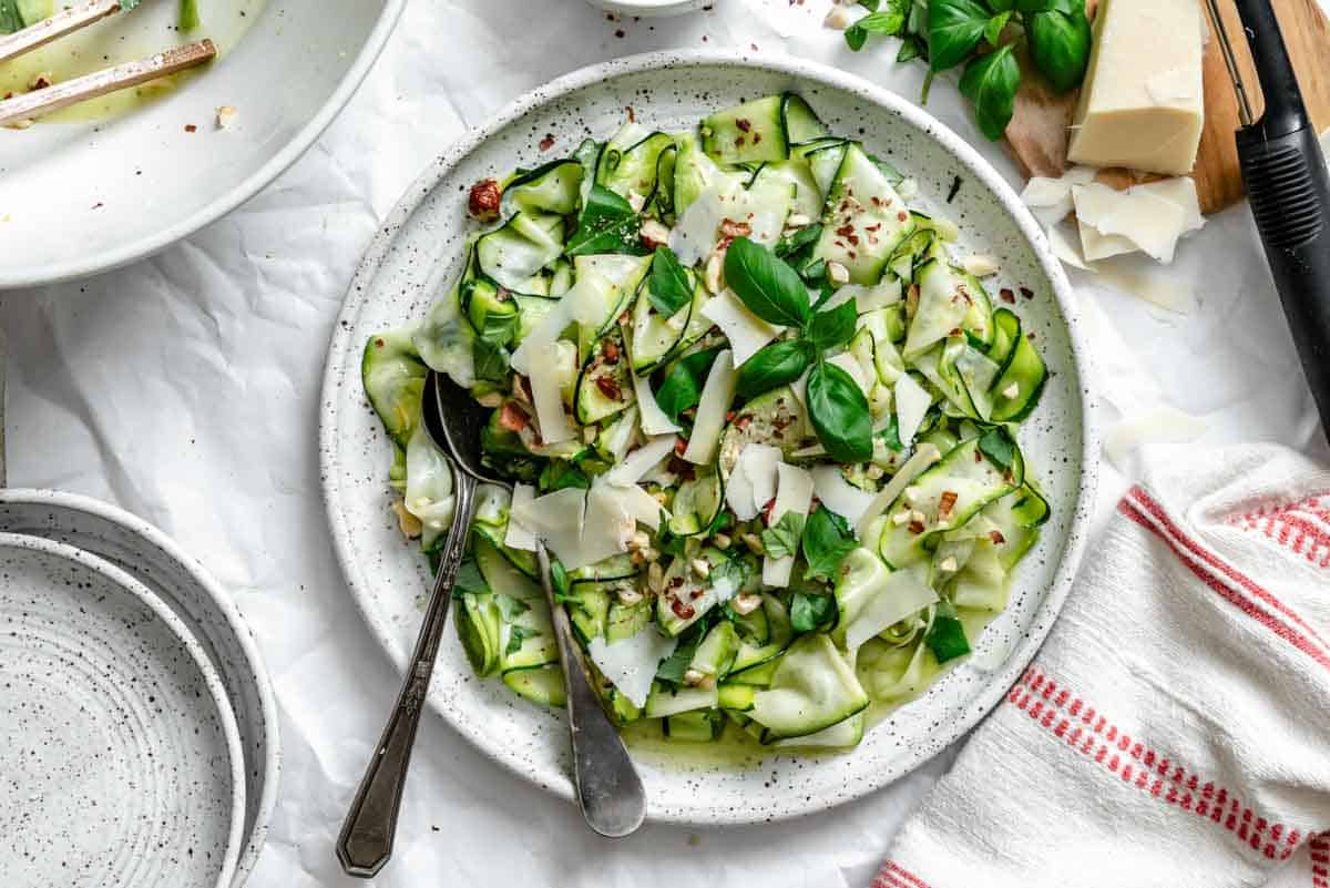 Overhead closeup of the completed zucchini ribbon salad on a white ceramic plate, showcasing the curled green zucchini ribbons intertwined with shaved parmesan, toasted hazelnuts, and vibrant basil leaves, with serving utensils.