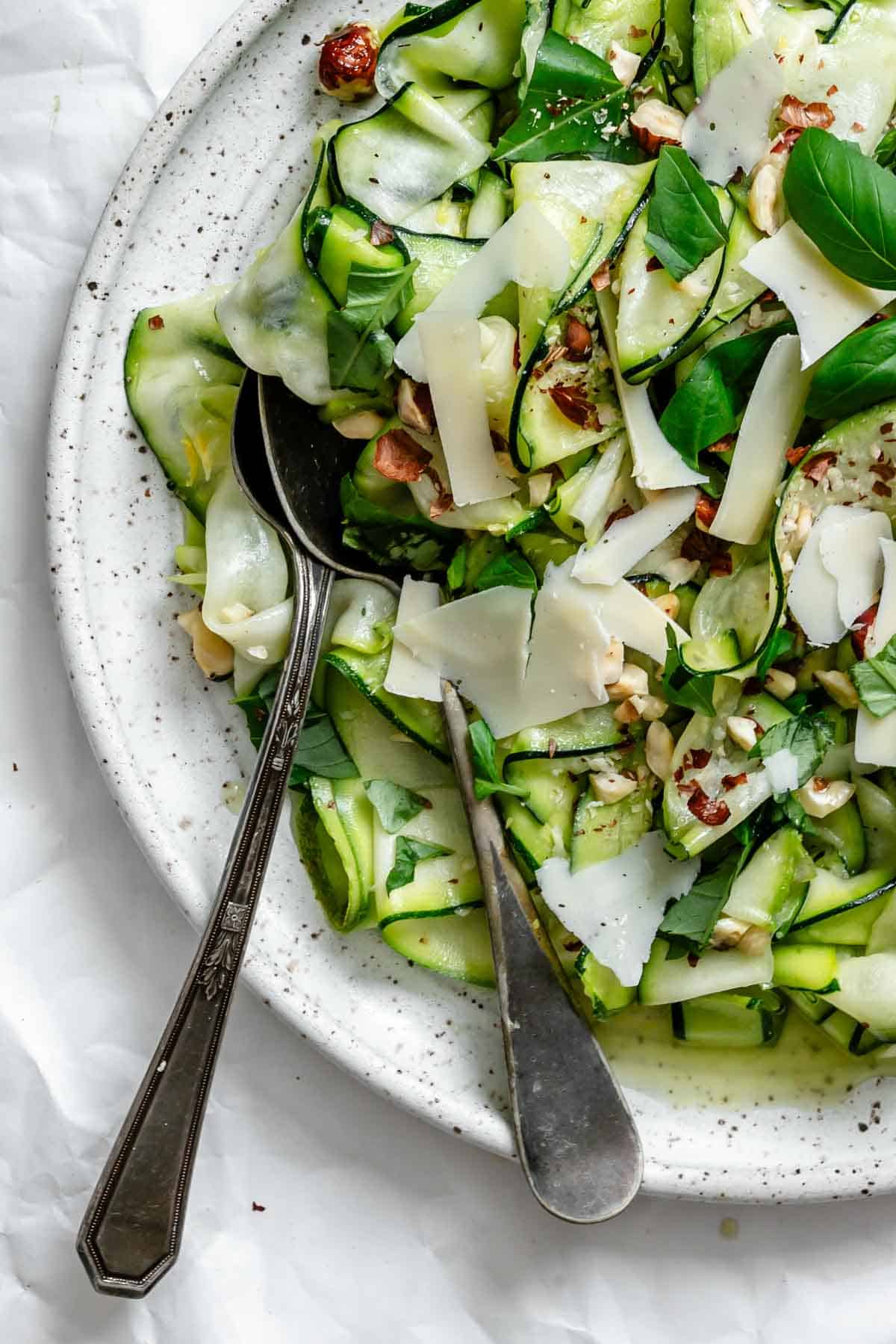 Extreme close-up of the salad highlighting the delicate texture of the zucchini ribbons, glossy from the dressing, with visible flecks of crushed red pepper, toasted hazelnuts, and fresh herbs tucked throughout the layers.