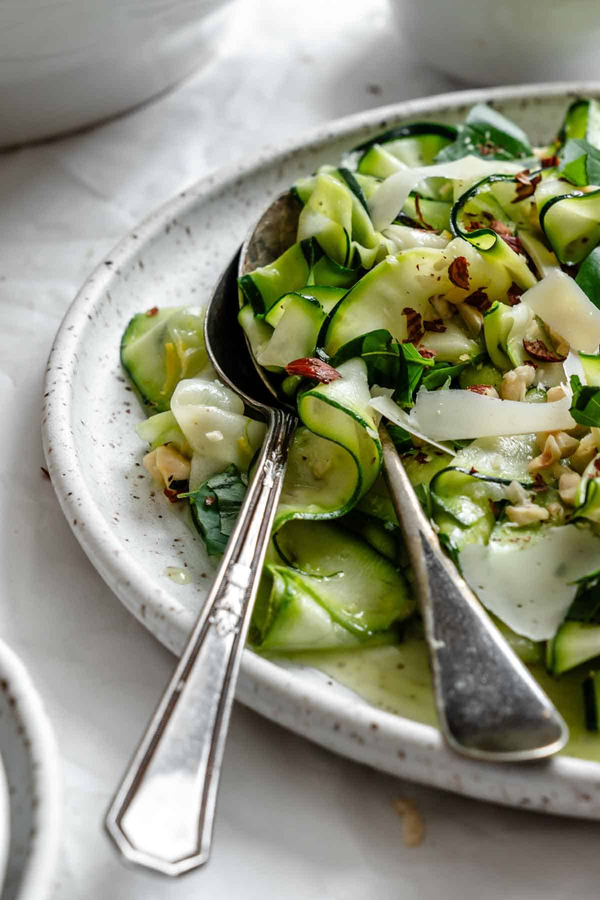 Artistic side angle of the zucchini salad with fork and knife placed in the dish, emphasizing the lightness of the ribbons and the contrast between the pale green zucchini, white parmesan shavings, and dark speckles of the ceramic plate.