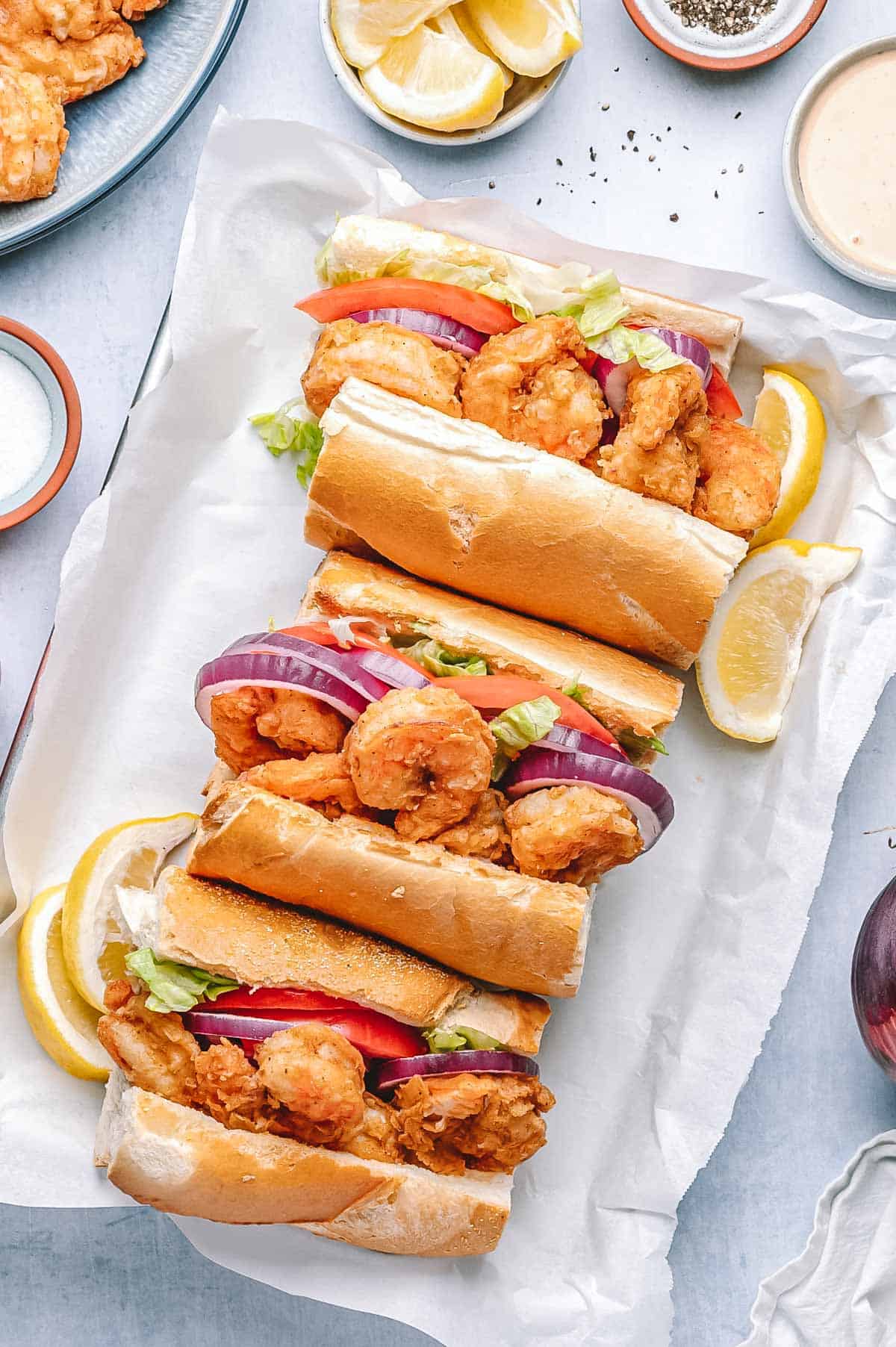 Overhead view of three shrimp po’boy sandwiches stuffed with crispy shrimp, sliced tomato, lettuce, and red onion on toasted French bread, garnished with lemon wedges and served with sauce.