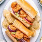Shrimp po’boys served on a white plate with crispy potato chips. Sandwiches are filled with fried shrimp, tomatoes, lettuce, and red onion, with lemon wedges and creamy sauce on the side.
