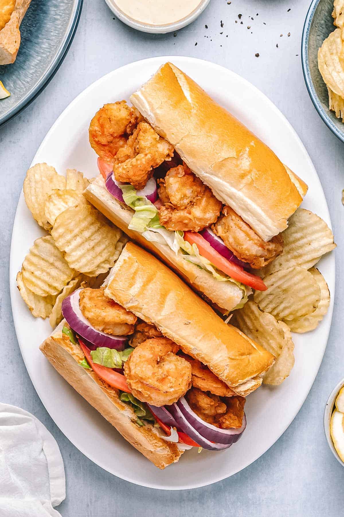 Shrimp po’boys served on a white plate with crispy potato chips. Sandwiches are filled with fried shrimp, tomatoes, lettuce, and red onion, with lemon wedges and creamy sauce on the side.